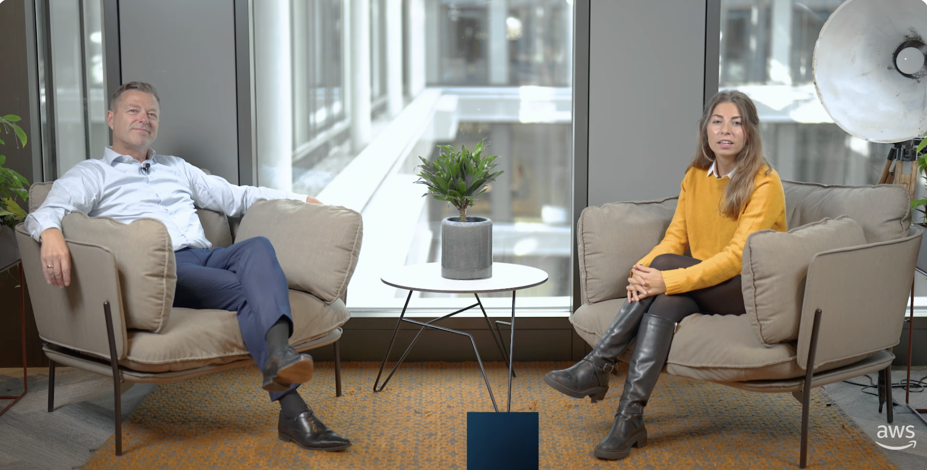 Two people sitting in armchairs having an interview in a modern office setting with large windows, a small table with a plant, and the AWS logo visible.