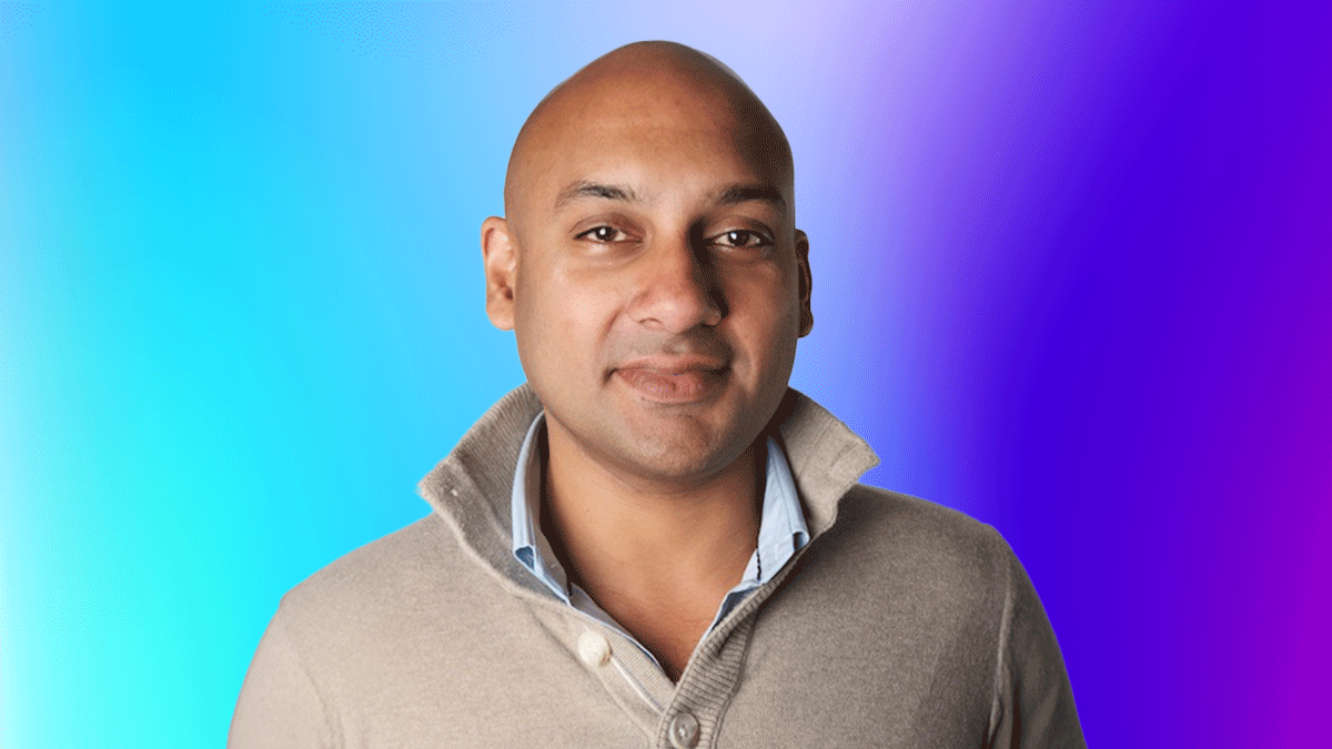 A portrait of a person with a shaved head wearing a beige sweater, standing in front of a blue and purple gradient background.