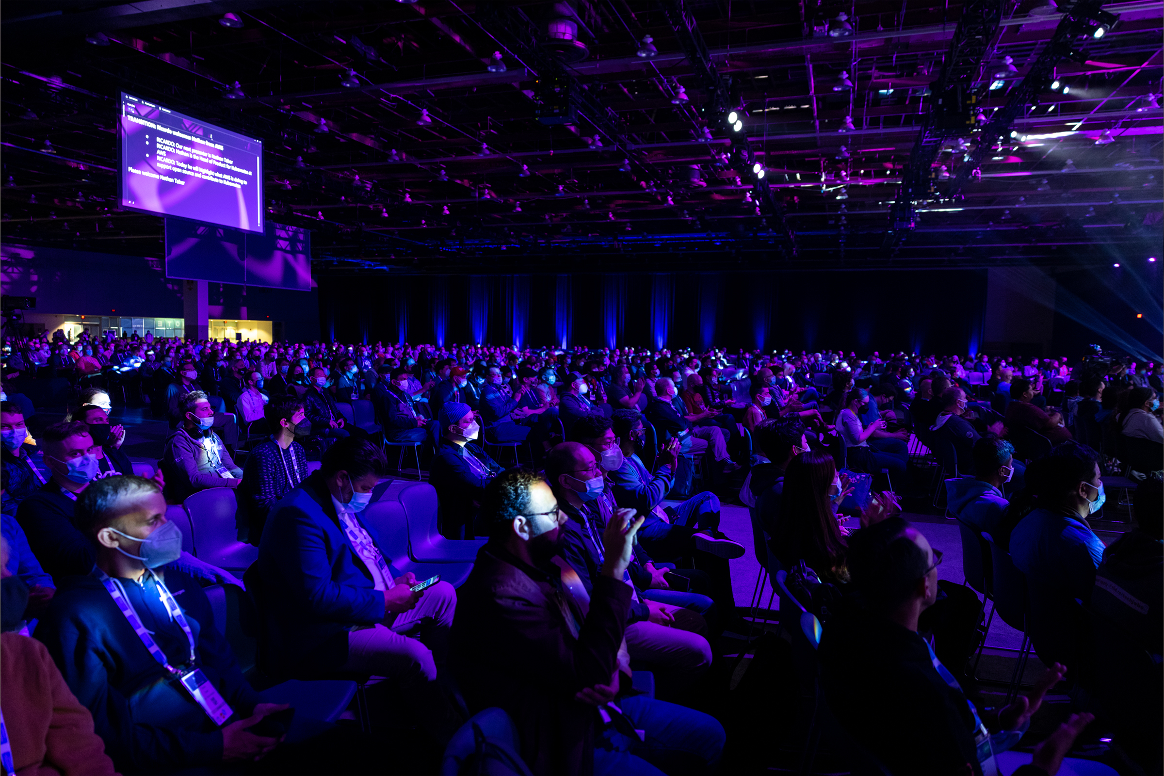 A large audience attending an AWS Retail KubeCon event in a conference hall, illuminated by purple and blue lighting. The attendees are seated and appear to be listening to a presentation, with many wearing face masks for safety.