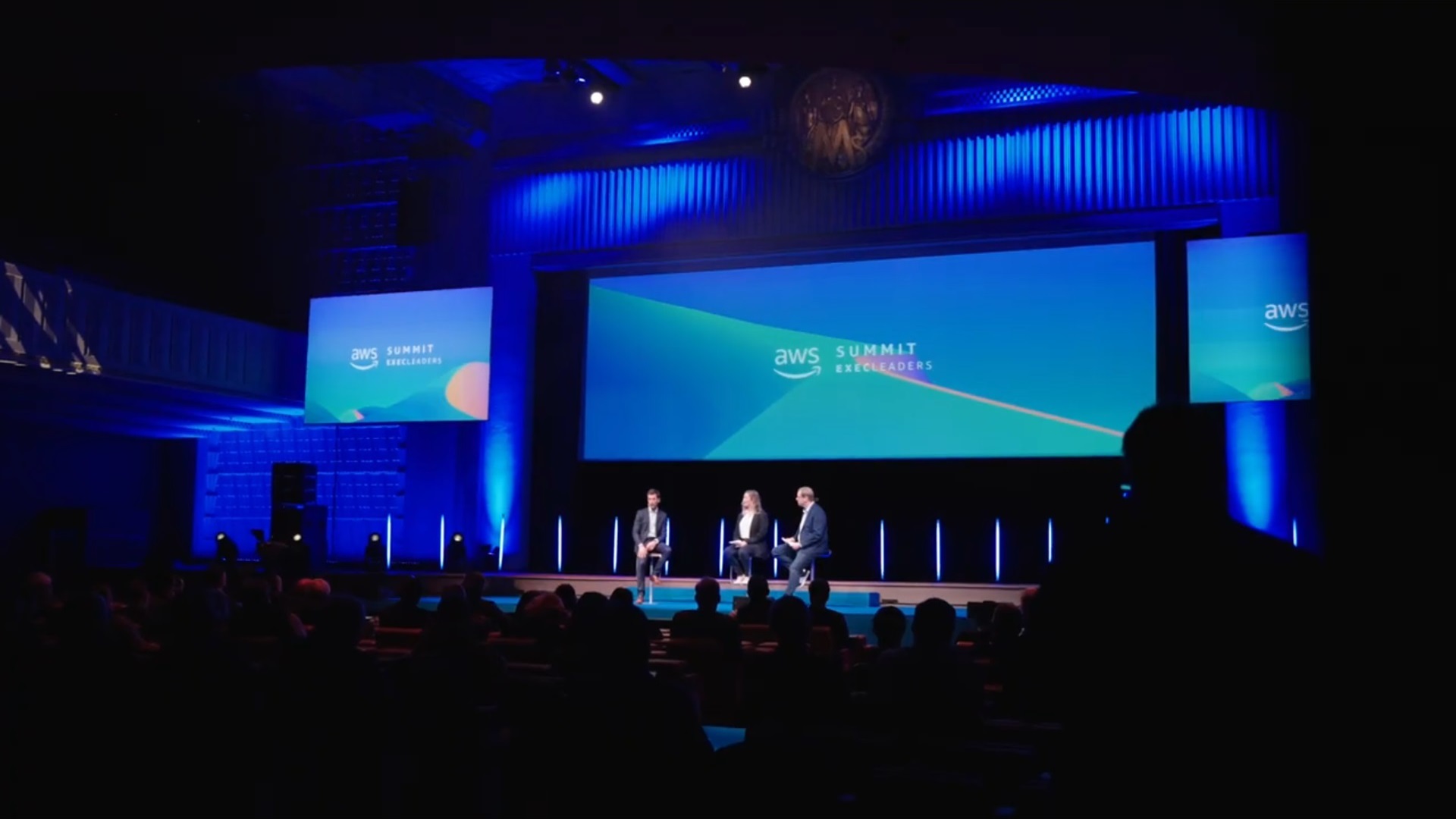 A panel discussion at the AWS Summit ExecLeaders event, showing three speakers on stage in front of an audience, with blue stage lighting and a large screen displaying the AWS Summit ExecLeaders branding.