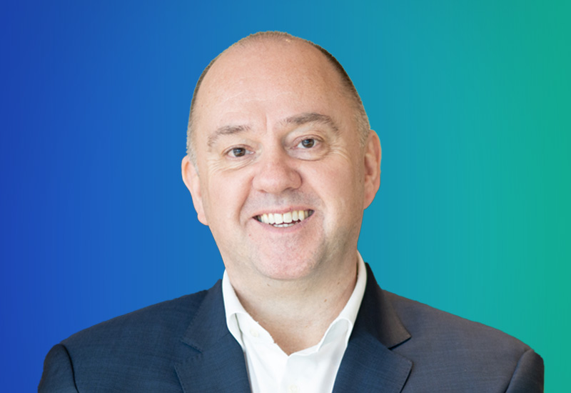 A professional executive headshot of a smiling person in a suit, posed in front of a blue and green gradient background.
