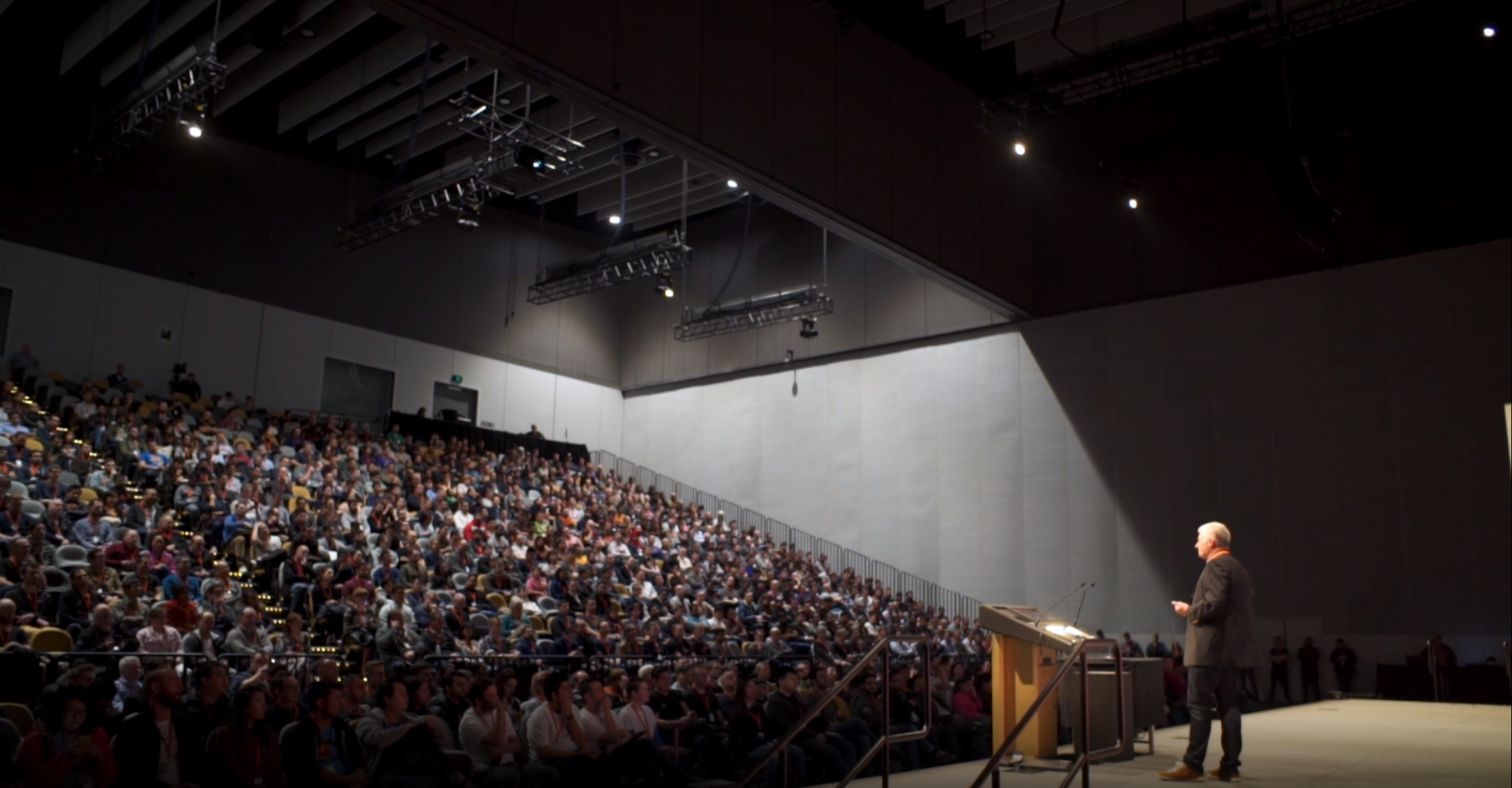 A speaker stands on a brightly lit stage addressing a large seated audience in a spacious auditorium.