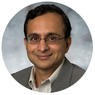 2A person wearing a gray suit jacket and a checkered shirt against a neutral background.