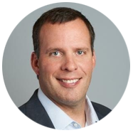 2A person wearing a dark suit jacket and white collared shirt against a neutral background.