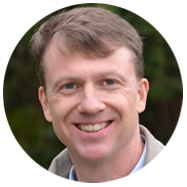 2A person with short brown hair wearing a light-colored jacket, set against a blurred green outdoor background.