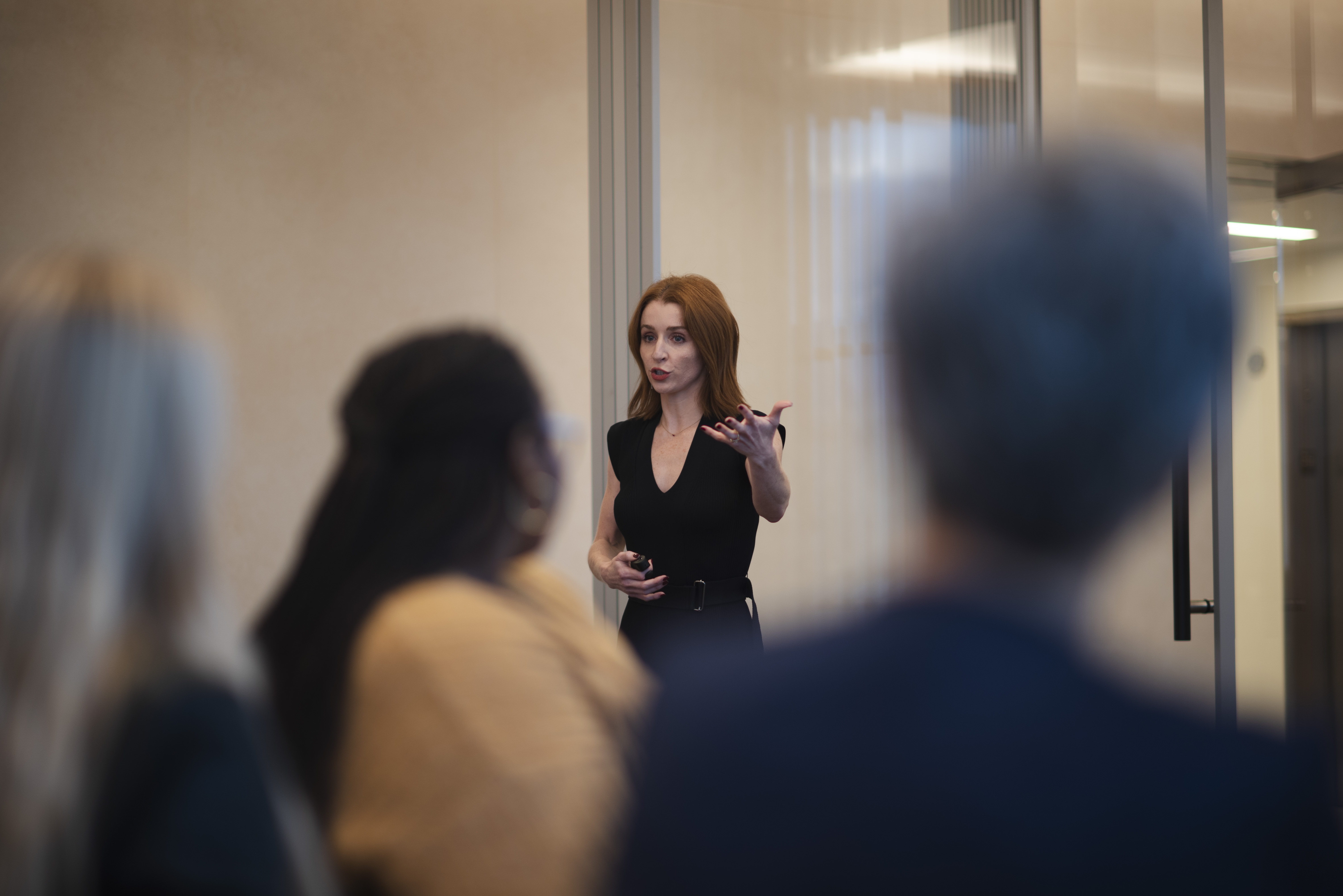 A woman in a black dress gestures while speaking to an audience in a modern indoor setting.