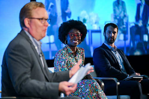 Three speakers participate in a panel discussion on stage at an AWS event, with a blue-lit background and audience in attendance.