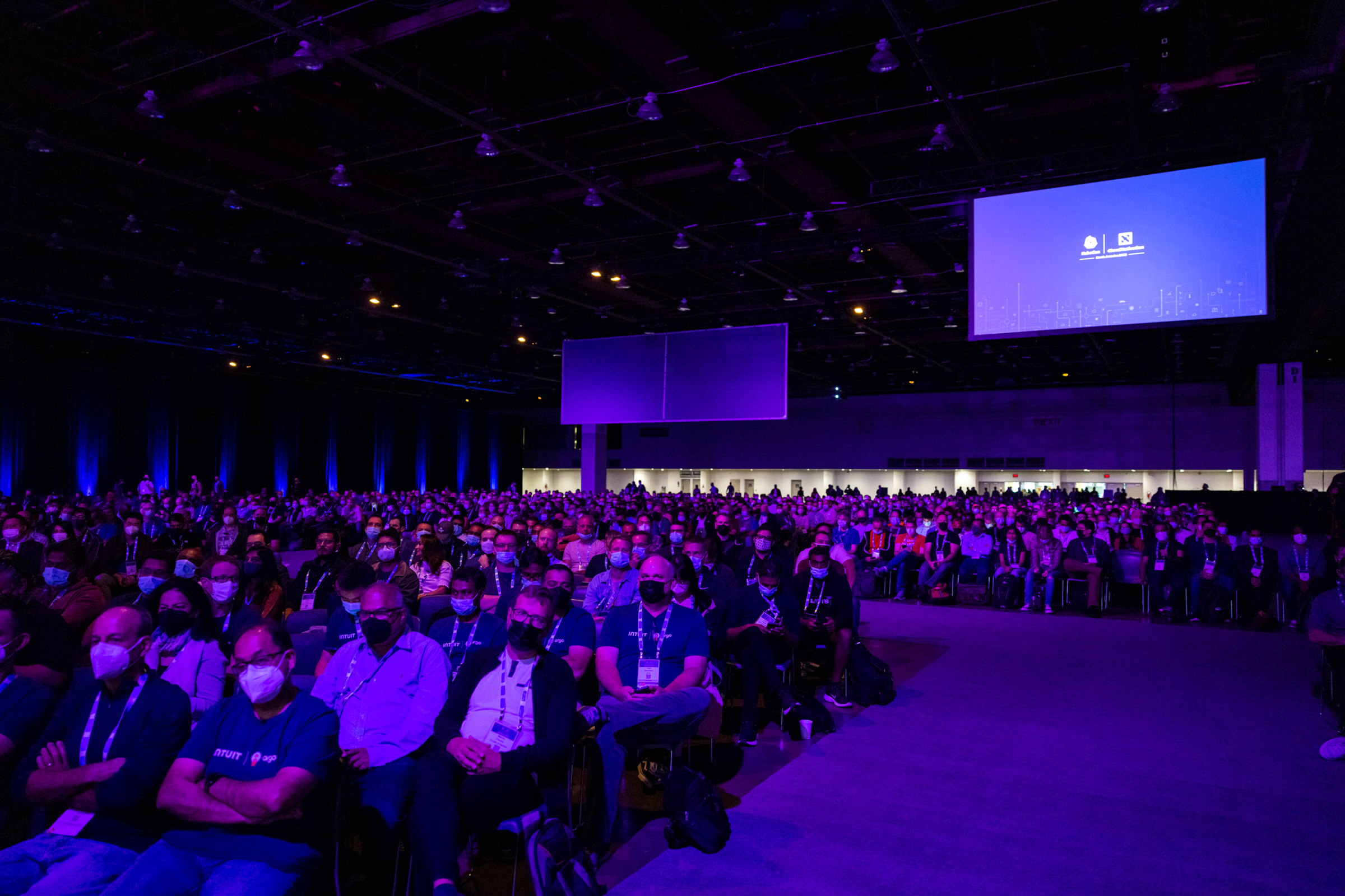 A large audience seated at an AWS Retail KubeCon event, illuminated by blue and purple stage lighting, with many attendees wearing masks. The scene captures the scale and engagement of a technology conference.