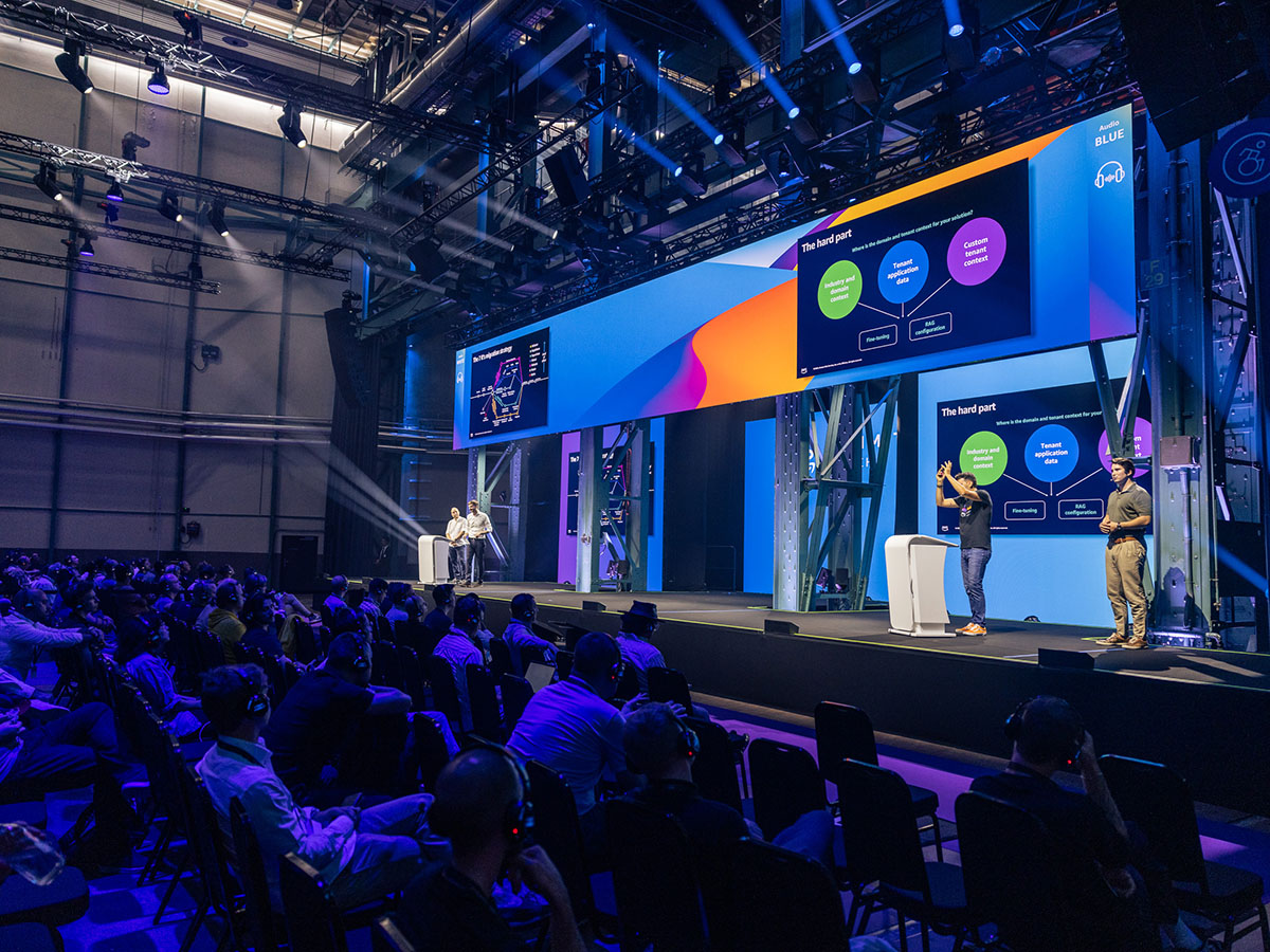 A large conference room with a seated audience watching presenters on stage under bright lights, with colorful slides displayed on a wide screen.