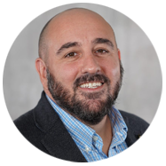 2A person wearing a blue checkered shirt and a dark blazer, set against a neutral gray background.