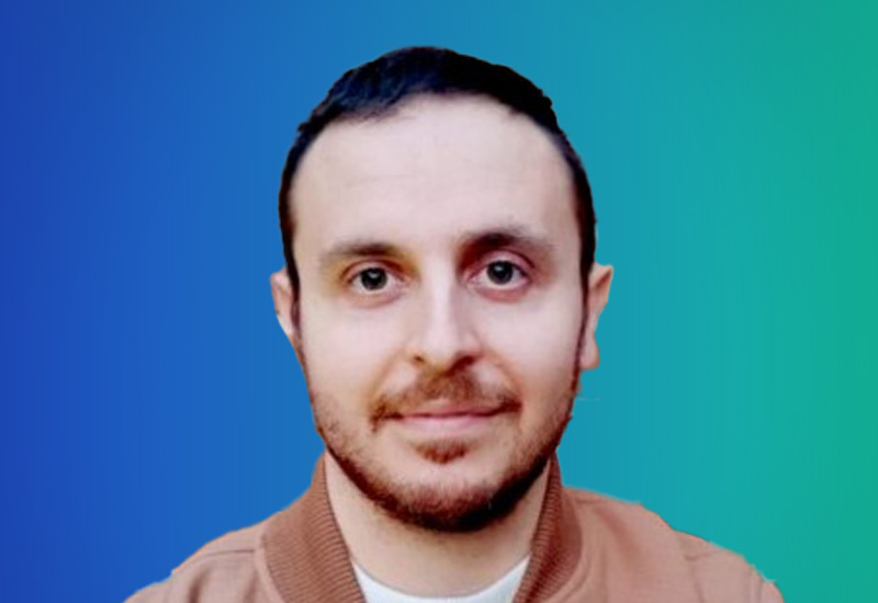 A headshot photo of a person with short dark hair and a trimmed beard, wearing a light brown jacket, against a blue and green gradient background.