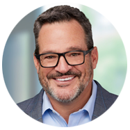 2A person wearing a blue collared shirt and a gray blazer, with a blurred background.