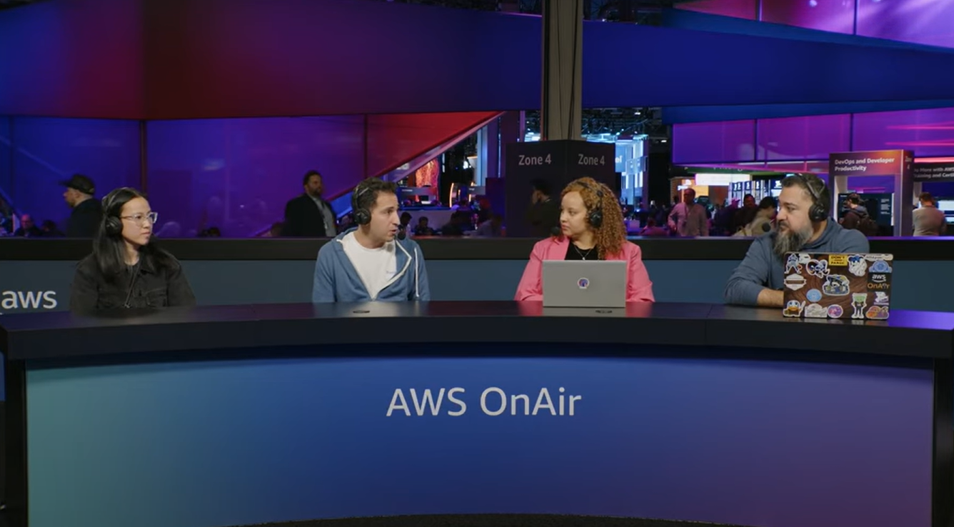 Four people participate in a live AWS OnAir panel discussion event at an AWS conference, seated at a desk with laptops and microphones, with a vibrant event floor and signage in the background.
