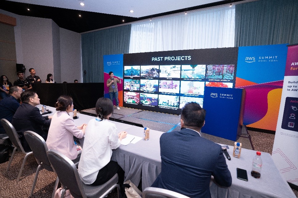 A presenter delivers a startup pitch in front of a panel of judges at the AWS Summit Hong Kong, with a display showing past projects in the background.