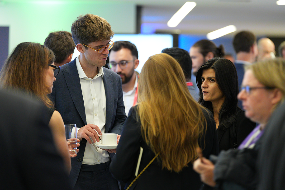 A large group of people in business attire networking at an indoor event, with one person holding a cup and saucer while talking.
