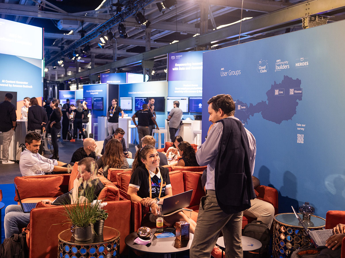 A busy tech event with attendees seated on red couches and others engaging at booths displaying technology-related content, set against a backdrop featuring a map and "AWS User Groups" branding.