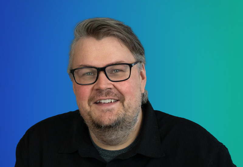 A professional headshot of a person with glasses and a beard, smiling in front of a blue and green gradient background.