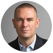 2A person wearing a dark suit jacket and light blue collared shirt, set against a gray background.