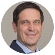 2A person wearing a suit jacket and a light blue collared shirt, set against a neutral background.