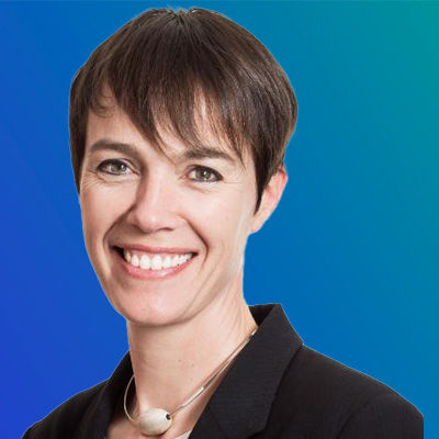 A person with short dark hair wearing a black blazer and a silver necklace, set against a blue gradient background.