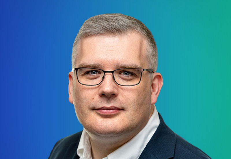 A person wearing a dark blazer and white shirt against a gradient blue-to-green background.