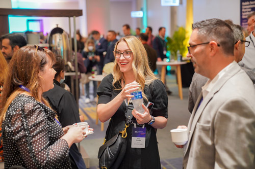 A group of professionals networking and conversing at an AWS event, with one woman holding a water bottle and others holding coffee cups in a lively conference atmosphere.