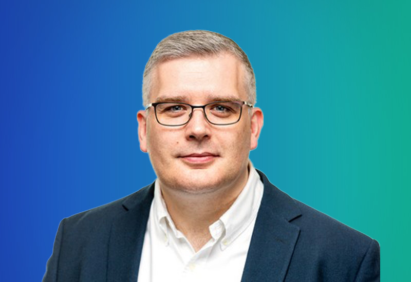 A professional headshot of a person wearing glasses, a white shirt, and a dark blazer, posed against a blue-green gradient background.