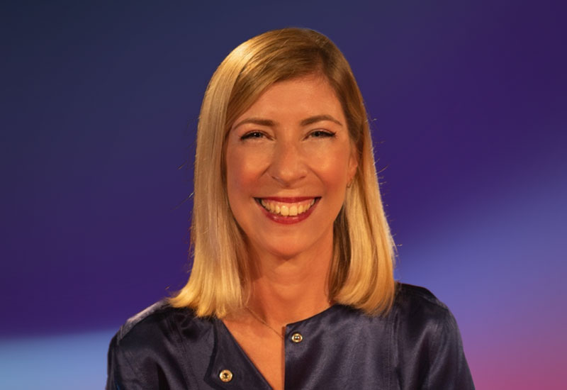 A person with shoulder-length blonde hair wearing a dark blue satin blouse, set against a gradient purple background.