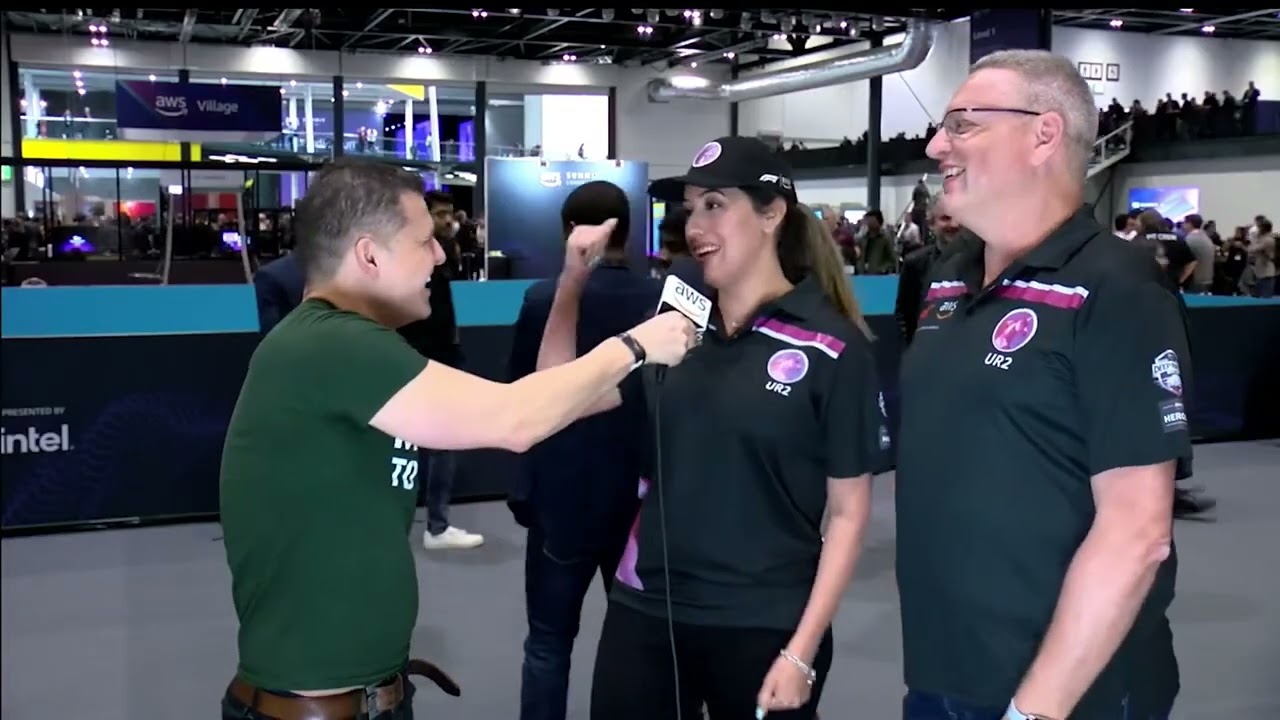 A scene from an AWS technology event showing an interviewer holding a microphone and speaking with two attendees. The background features the AWS Village and a busy expo floor with people and booths.