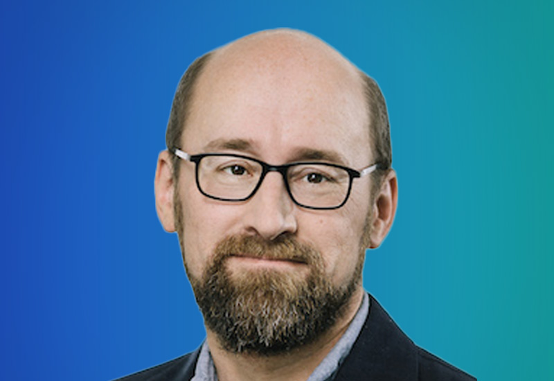 A person with a bald head and short hair on the sides, wearing a dark jacket against a gradient blue-green background.