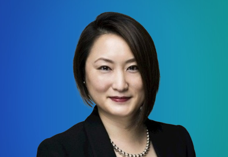 A person wearing a black blazer and a pearl necklace, set against a blue-to-teal gradient background.
