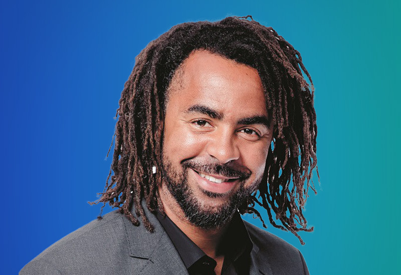 A person with long, dark dreadlocks wearing a gray suit and black shirt, set against a blue gradient background.