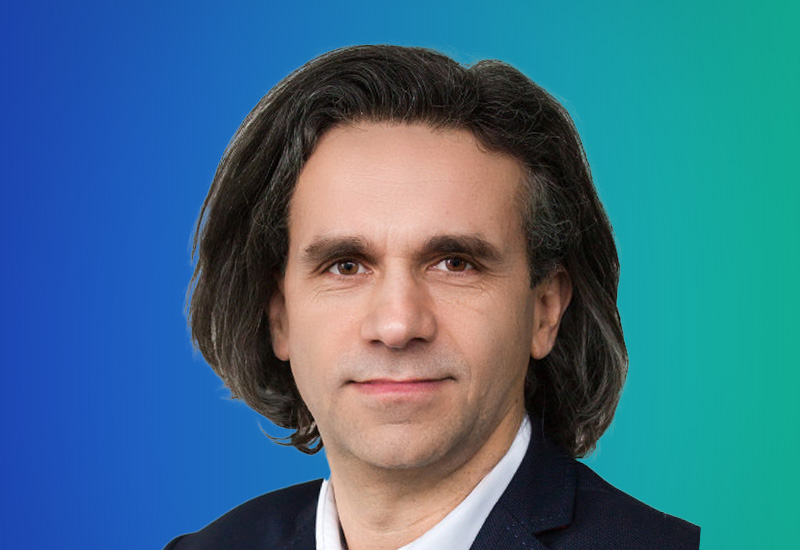 A person with shoulder-length dark hair wearing a suit against a gradient blue and green background.