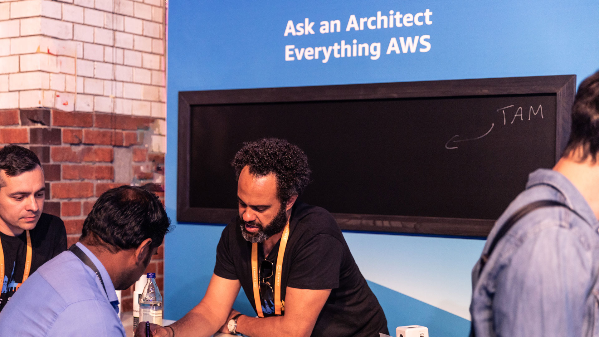 A booth with a sign reading "Ask an Architect Everything AWS," featuring a chalkboard labeled "TAM" and people engaged in discussion.
