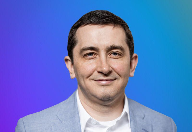 A person wearing a light blue blazer and white shirt against a gradient blue and purple background.