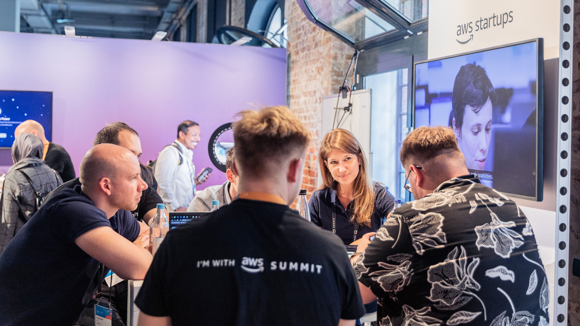 A group of people gathered around a table at an AWS Startups booth, with a screen and branding visible in the background.