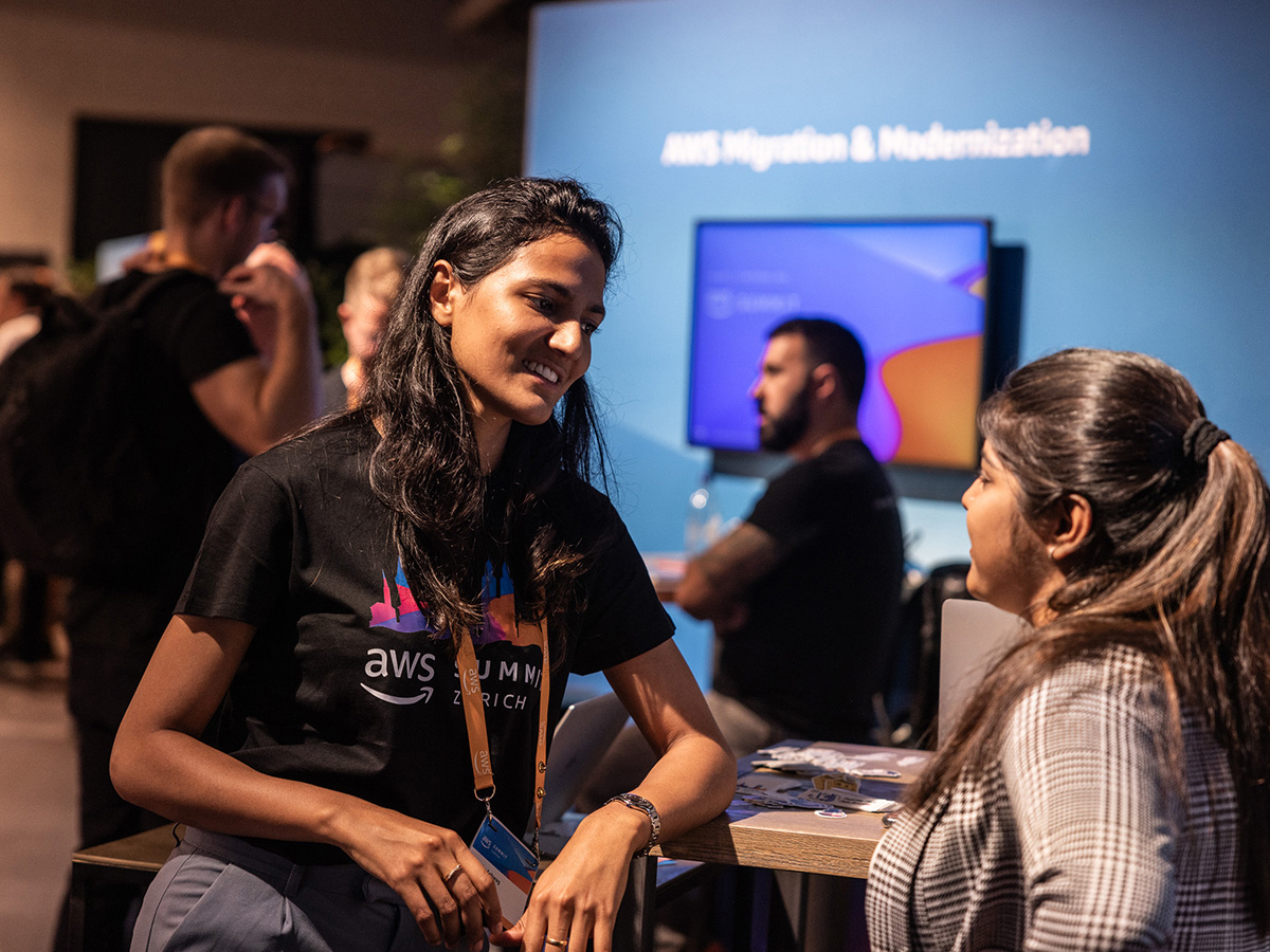 People networking at an AWS Summit booth with a screen displaying "AWS Migration & Modernization" in the background.