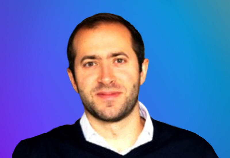 A portrait of a person with short dark hair and a trimmed beard, set against a vibrant blue and purple gradient background.