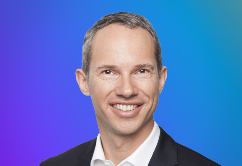 A person in formal attire against a gradient blue and purple background.