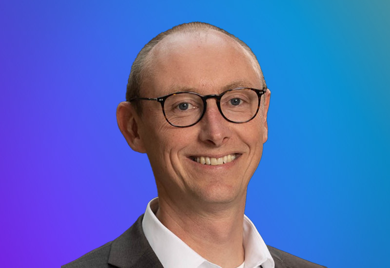 Professional headshot of a smiling person with glasses, wearing a suit and white shirt, set against a blue and purple gradient background. Ideal for Stockholm Summit event materials and profiles.