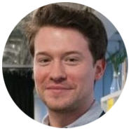 2A person wearing a white collared shirt in an indoor setting with shelves and decor in the background.