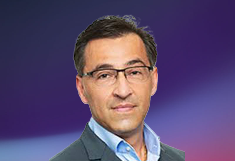 A person wearing a suit and light blue shirt against a gradient purple background.