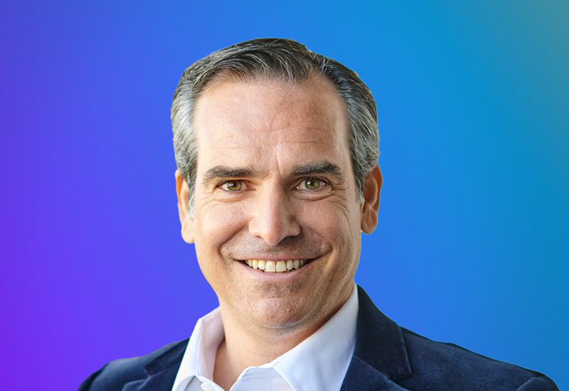 Portrait headshot of a smiling business professional in a suit jacket with an open-collar shirt, set against a vibrant blue and purple gradient background. Suitable for AWS Summit Paris event materials or speaker profiles.
