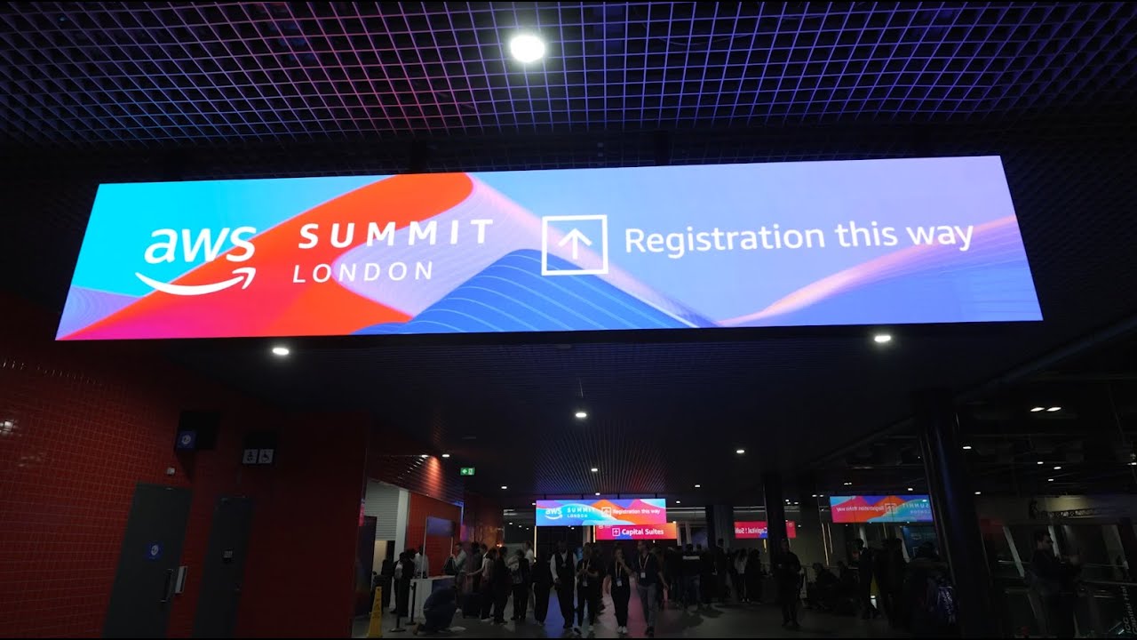 "Sign reading 'AWS Summit London - Registration this way' with an arrow, above a crowd in a conference venue."