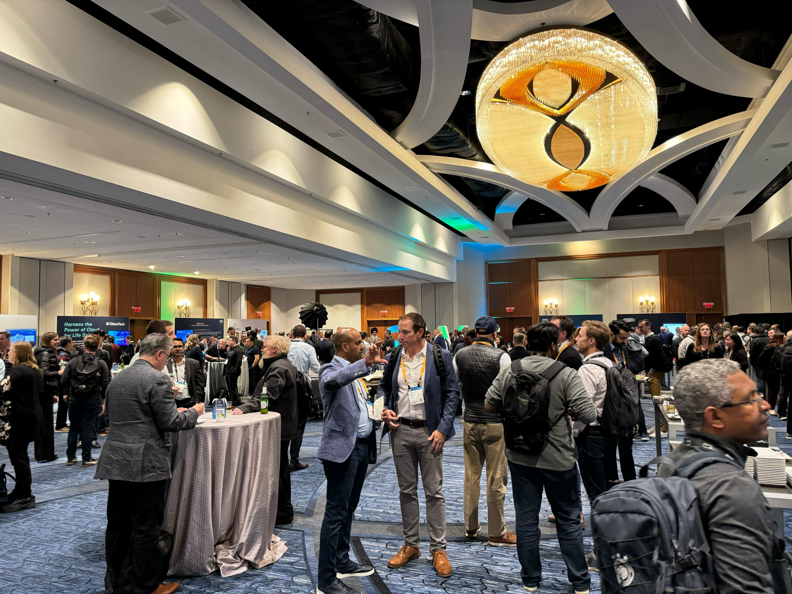 A networking event in a large conference hall with groups of people conversing, a round table with drinks, and a prominent chandelier overhead.
