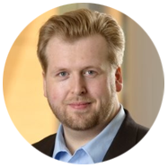 2A person wearing a suit jacket and light blue collared shirt, with a blurred background.