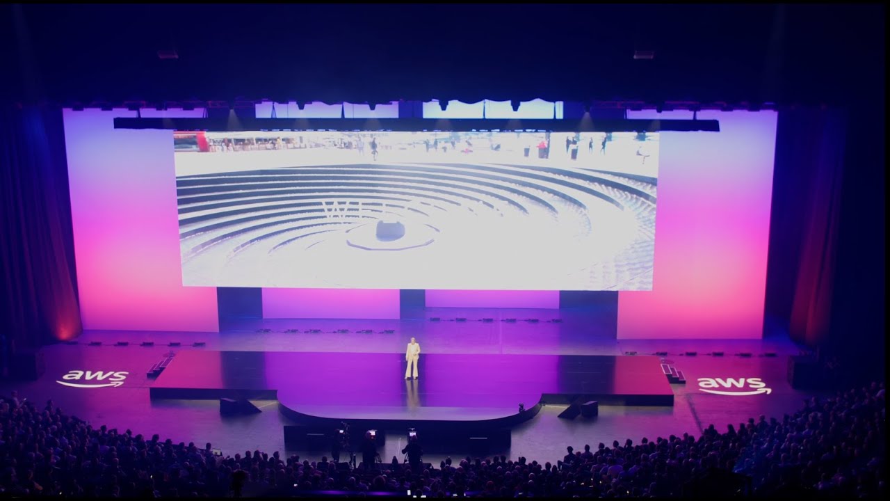A large stage with purple lighting, a speaker standing in front of a crowd, and a screen displaying an image of circular steps; AWS branding visible on the stage.