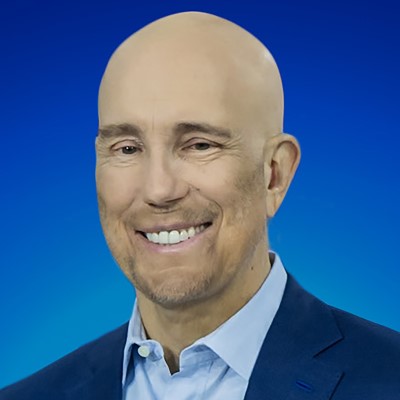A professional headshot of a smiling, bald man in a blue suit and light blue shirt against a blue gradient background.