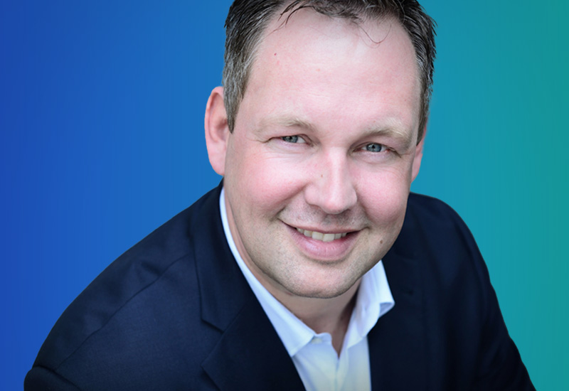A person wearing a dark suit and white shirt against a gradient blue-green background.
