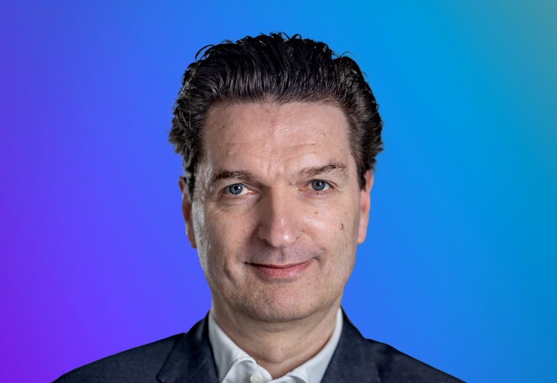 A person wearing a suit and white shirt against a gradient blue and purple background.
